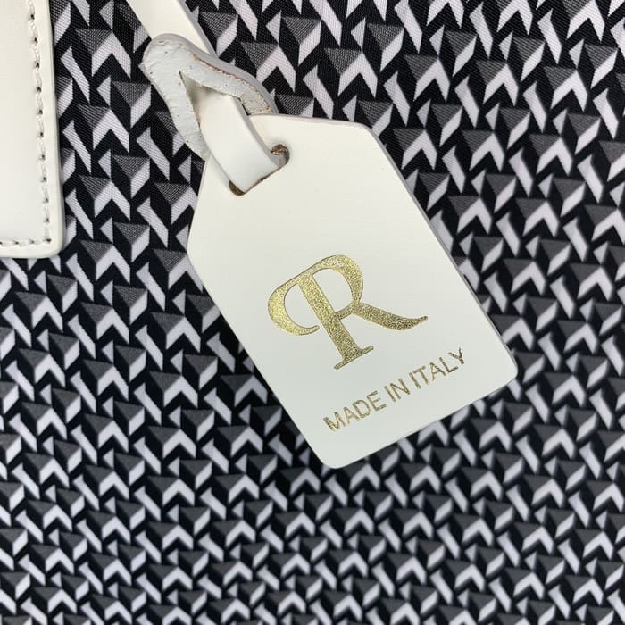 흑백 기하학적 패브릭의 배경에는 금색으로 된 문자 "R"과 "MADE IN ITALY"라는 단어가 새겨진 흰색 가죽 ROBERTA PIERI 태그가 걸려 있습니다.