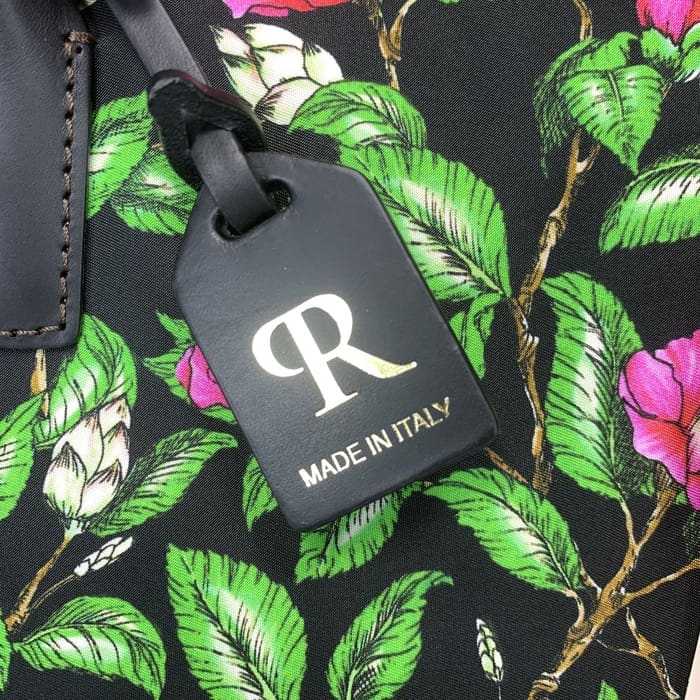 골드 "R"과 이탈리아 텍스트가 새겨진 블랙 ROBERTA PIERI 러기지 태그는 녹색 잎과 분홍색 꽃이 있는 꽃무늬 패브릭에 매달려 시대를 초월한 이탈리아 우아함으로 미니 더플을 보완합니다.