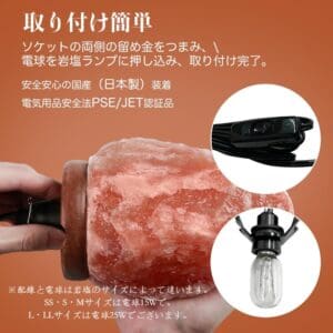 手が「天然岩塩ランプ【サイズ：SS】—ほっと落ち着く、やさしい間接照明」に電球を差し込み、お部屋の間接照明としてその穏やかな自然な輝きを際立たせています。 日本語の説明文には、ランプの安全機能、公式認証、および対応する電球サイズが記載されています。上部にはスイッチ付きの黒いコードが写っており、このヒマラヤ岩塩アクセサリーの利便性と実用性を際立たせています。.