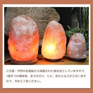 提供された製品データを反映して修正した文章は以下の通りです：3つの天然岩塩ランプ【サイズ：SS】—ほっと落ち着く、やさしい間接照明—が木製の台の上に置かれ、その優しい光で部屋を柔らかく照らし、エレガントな間接照明となっています。 以下の日本語の注記には、天然素材であるため、ランプごとに外観が自然に異なることが説明されています。.